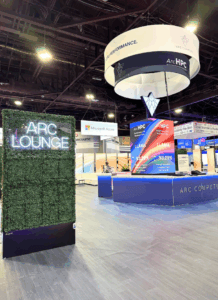 Arc Compute custom booth with a large hanging sign, lounge, custom counter and a center tower featuring LED video tiles.